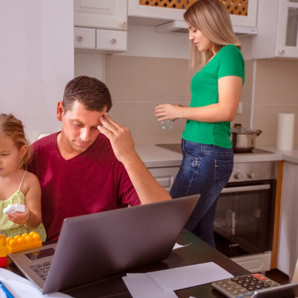 Stressed man working on a laptop at the kitchen table, surrounded by bills and paperwork, while a young child plays nearby and a woman prepares a meal in the background. Overwhelmed with family and financial responsibilities.