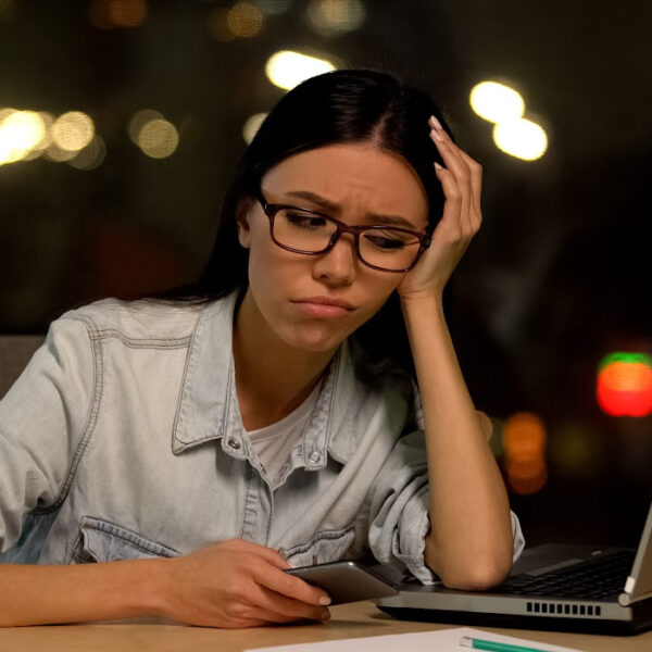 Frustrated woman holding a smartphone while looking at her laptop, appearing mentally drained and overwhelmed. Concept of time management struggles, task switching, and lost productivity.