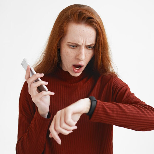 Shocked woman checking the time on her smartwatch while holding a smartphone, looking frustrated or surprised about being late. Concept of time management, running late, or losing track of time.