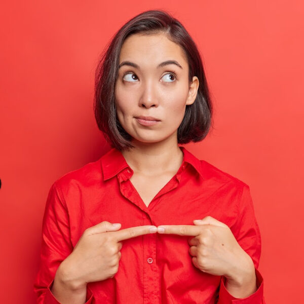 Woman in red shirt pointing at herself with a puzzled expression against a red background—representing self-awareness and mindset shifts tied to daily habits that feel productive but keep you stuck.